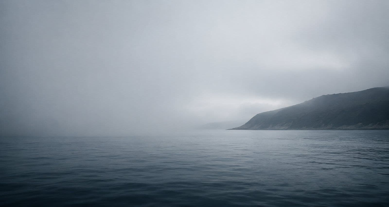 A wide expanse of calm water as fog begins to lift, a low dark headland barely resolving on the right and the horizon still unresolved.