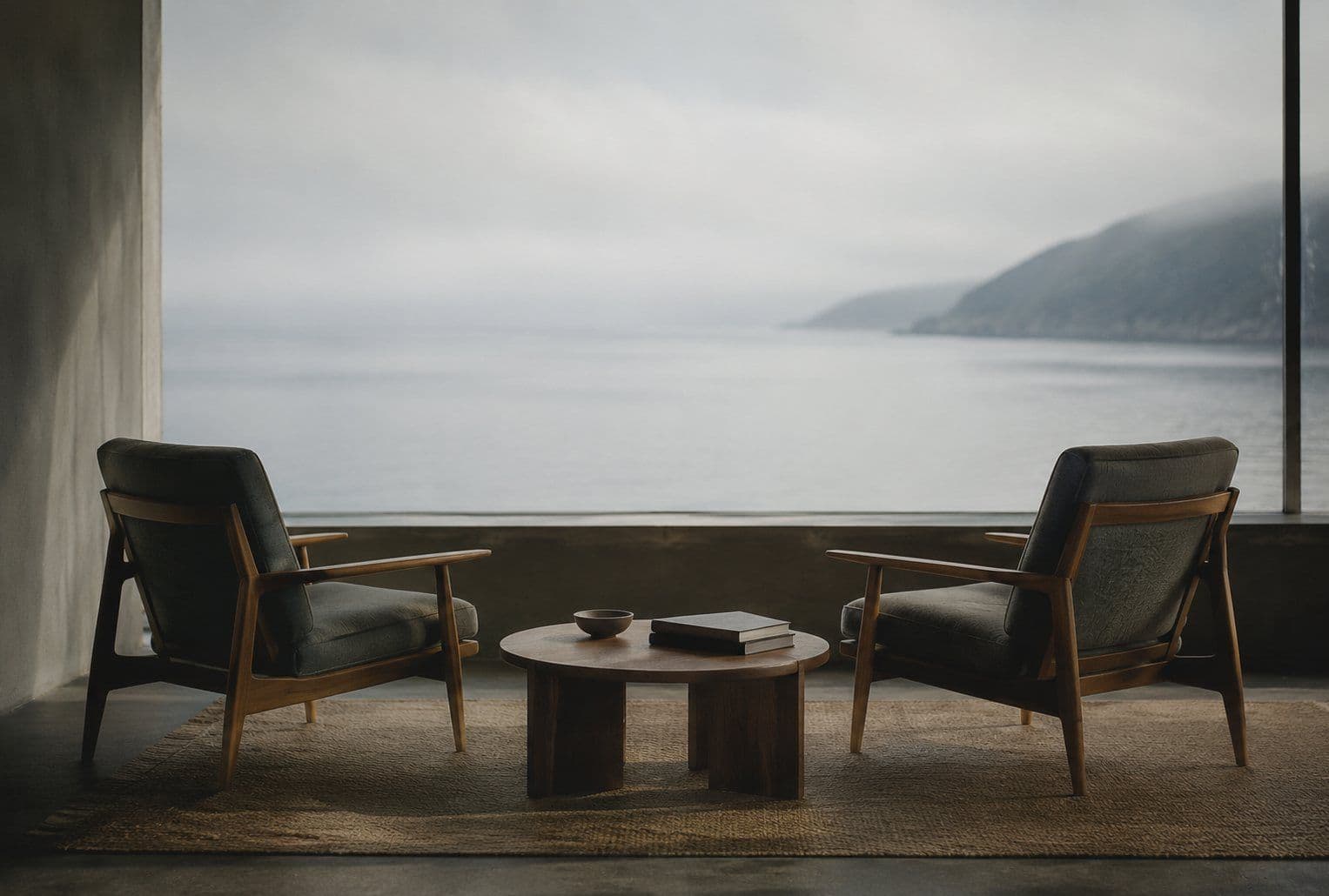 Two simple wooden chairs angled toward one another beside a tall window, a small low table between them with a closed notebook on it.